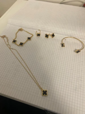 Set med fyrklöver smycken i guld - Säljer ett matchande smyckeset med halsband, armband, örhängen och armring i guldfärg med svarta fyrklöverformade hängen. Alla delar har en elegant och modern design med guldfärgad metall och svart emalj. Perfekt för dig som gillar stilrena accessoarer med en twist.