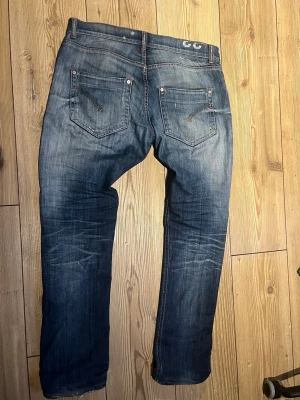 Dondup jeans - Säljer ett par blå jeans med slitningar och hål på båda benen. Jeansen har fem fickor, dragkedja och knapp i midjan samt detaljerade sömmar på bakfickorna. Perfekta för en avslappnad och trendig streetstil.