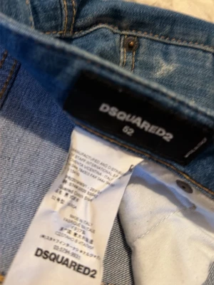 Blå jeansshorts från Dsquared2 - Säljer ett par blå jeansshorts från Dsquared2 med slitna detaljer och färgstänk. Shortsen har två bakfickor och en unik patch med färgglatt tryck. Klassisk denimlook med synliga sömmar och knappgylf.