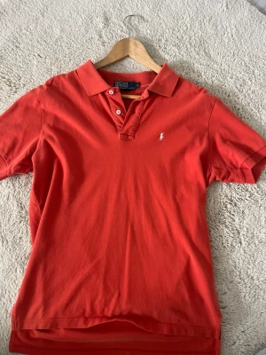 Orange pikétröja från Polo Ralph Lauren - Klassisk orange pikétröja från Polo Ralph Lauren med korta ärmar och vit broderad logga på bröstet. Tröjan har krage, knappar vid halsen och är gjord i mjuk bomull. Perfekt till en avslappnad men ändå stilren look.