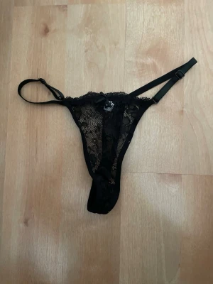 Svart spetsstring med rosett - Säljer en svart stringtrosa i genomskinlig spets med blommigt mönster och liten rosett framtill. Trosan har tunna, justerbara band i sidorna och en sexig, smal passform. Perfekt för dig som gillar detaljer och en stilren look.Samfraktar.
