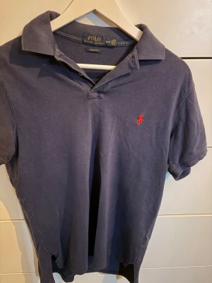 Marinblå pikétröja från Ralph Lauren - Klassisk marinblå pikétröja från Polo Ralph Lauren med custom fit och korta ärmar. Tröjan har den ikoniska röda logobroderingen på bröstet och mjuk krage. Perfekt till jeans eller shorts för en clean look.