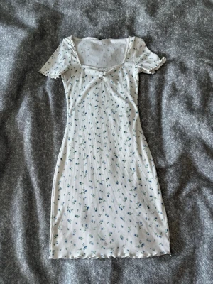 Vit blommig kortklänning - Söt vit kortklänning med små blå blommor och ribbad struktur. Klänningen har korta ärmar, lätt v-ringad hals och volangdetaljer vid ärmslut och nederkant. Perfekt för varma dagar och ger en romantisk vibe.