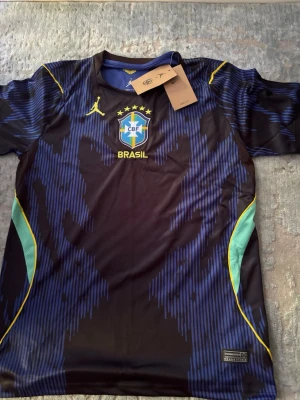 Brasil fotbollströja Nike x Jordan - Snygg svart och blå Brasilien fotbollströja från Nike x Jordan med CBF-emblem och fem stjärnor broderade på bröstet. Tröjan har gula detaljer, rund hals och är tillverkad i lätt, snabbtorkande Dri-FIT-material. Perfekt för träning eller match.