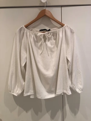 Vit offshoulder blus med knytband - Trendig vit offshoulder blus med långa ärmar och knytband framtill. Tillverkad i ett lätt, ribbat tyg som ger en avslappnad och luftig känsla. Perfekt att styla med jeans eller kjol för en cool och enkel look.