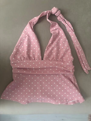 Rosa prickig halterneck topp - Supersöt rosa halterneck topp med vita prickar och knyt i nacken. Toppen har en djup v-ringning och är ärmlös, perfekt för varma dagar. Materialet känns mjukt och stretchigt, troligen polyester eller bomullsmix. Passar dig som vill ha en lekfull och trendig look.
