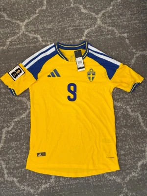 Svensk landslagströja Adidas gul - Gul svensk landslagströja från Adidas med blå detaljer och nummer 9 på bröstet. Tröjan har blå och vita ränder på axlarna, FIFA-märke på ärmen och det svenska landslagets emblem på bröstet. Tillverkad i lätt och ventilerande material.