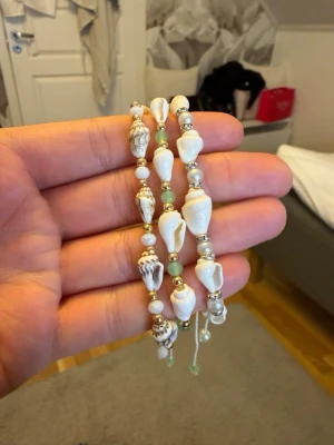 Armband med snäckor och pärlor - Snyggt armband med vita och bruna snäckor, små vita pärlor och gröna samt guldfärgade detaljer. Armbandet har en bohemisk känsla och passar perfekt för dig som gillar strandinspirerade accessoarer. Justerbar storlek med snöre för enkel användning.