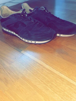 Svarta sneakers med nitar från Valentino - Snygga svarta sneakers från Valentino med coola silverfärgade nitar runt sulan. Skorna har en låg profil, svart mocka och läderdetaljer samt vita insidor. Perfekta för dig som vill ha en unik och trendig look. Passar riktigt fint på kvällarna eller om man ska på en dejt.