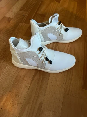 Vita sneakers med elastisk snörning - Stilrena vita sneakers med elastisk snörning och detaljer i beige mesh. Skorna har en modern, sportig look med högt skaft och mjuk sula. Materialmix av syntet och mesh ger både komfort och ventilation. Perfekta för dig som gillar en clean och fräsch stil.