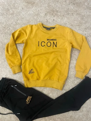 Gult och svart set ICON DSQUARED2 - Snyggt set med gul sweatshirt och svarta byxor från DSQUARED2. Tröjan har trycket 'IBRAHIMOVIC ICON' fram och en stor grafisk bild med DSQUARED2-logga bak. Byxorna har dragkedjefickor och matchande ICON-tryck. Perfekt för dig som gillar streetwear och statement-plagg.