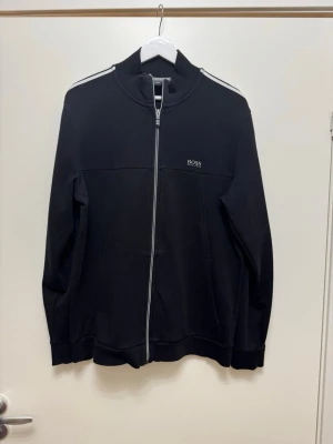 Svart zip tröja  från BOSS - Svart zip tröja från BOSS med hel dragkedja och vita ränder längs ärmarna. Liten BOSS-logga på bröstet och ståkrage. Tillverkad i mjukt material, perfekt för en stilren och sportig look.