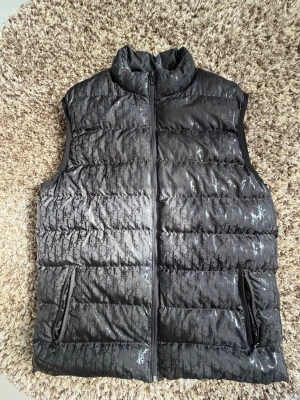 Grå dunväst med mönster från Dior - Snygg grå dunväst från Dior med ett diskret, upprepat logomönster över hela västen. Västen har quiltad design med vertikala sömmar och en glansig finish. Perfekt för dig som vill ha något både trendigt och bekvämt.