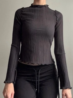 Svart topp - Trendig svart ribbad topp med lång ärm och lätt volangkant vid hals, ärmslut och nederkant. Figurnära passform och stretchigt material som ger en snygg siluett. Perfekt att styla med både jeans och kjol.