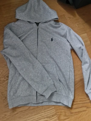 Grå hoodie från Polo Ralph Lauren - Snygg grå hoodie från Polo Ralph Lauren med dragkedja framtill och klassisk huva. Tröjan har det ikoniska lilla mörkblå logotrycket på bröstet och två fickor på sidorna. Tillverkad i mjukt bomullsmaterial som känns skönt mot huden. Perfekt för en avslappnad och stilren look.