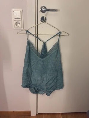 Blå spetsdetaljerad pyjamasbody - Söt blå pyjamasbody med tunna axelband som korsas i ryggen och spetsdetaljer längs urringningen. Luftigt material som känns mjukt mot huden, perfekt för chillkvällar hemma. Ingen synlig logga eller märke.