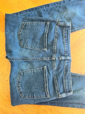 Blå bootcut jeans med stora bakfickor - Säljer ett par klassiska blå bootcut jeans med stora bakfickor och markerade sömmar. Jeansen har normalhög midja, knapp- och dragkedjestängning som går ut nedtill. Perfekta för en avslappnad och trendig look.