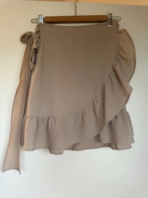 Beige omlottkjol med volang - Supersöt beige kort omlottkjol med volangkant och knytband i midjan. Kjolen har en flowy känsla och är perfekt för varma dagar. Tillverkad i ett lätt och mjukt material som känns skönt mot huden.
