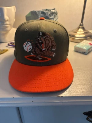 Grizzlies keps Oanvänd - Snygg keps i olivgrönt med orange skärm och broderad björn som slår en baseboll på framsidan. På sidan finns ett Grizzlies 20 Seasons-märke. Perfekt för dig som gillar sportig stil och vill sticka ut med cool brodyr.