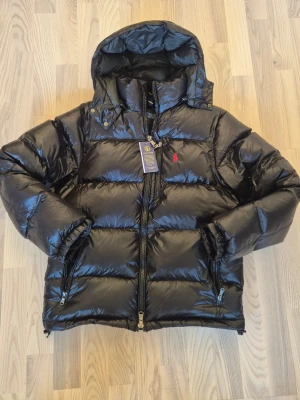Svart dunjacka från Polo Ralph Lauren - Snygg svart dunjacka från Polo Ralph Lauren med glansig finish, röd broderad logga på bröstet och avtagbar huva. Jackan har två dragkedjefickor. Jackan säljes för den var lite för liten. Personen i bilden är ca 175 och passar honom bra så om du är runt 170-180 så bör den passa berfekt