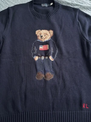 Ralph Lauren stickad tröja Polo Bear - Mörkblå stickad tröja från Ralph Lauren med den ikoniska Polo Bear på bröstet, klädd i jeans och tröja med amerikanska flaggan. RL-broderi i rött nere vid fållen. Perfekt nu i vintern och inför våren. Storlek L men passar M väldigt bra. Pris går att diskutera enbart vid snabbaffär.