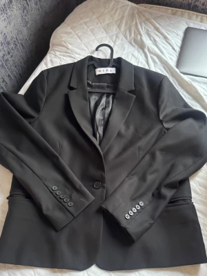 Svart kavaj från WERA - Stilren svart kavaj från WERA med klassisk passform, enkelknäppning och fyra knappar vid ärmsluten. Kavajen har två fickor framtill och är helfodrad för en snygg siluett. Perfekt för en trendig och uppklädd look.