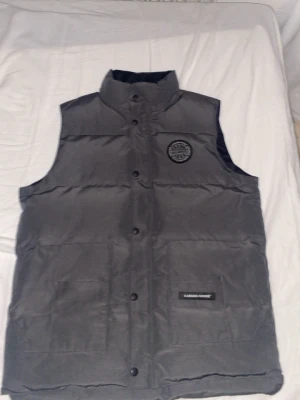 Grå dunväst från Canada Goose - Snygg grå dunväst från Canada Goose med hög krage och tryckknappar framtill. Västen har ett broderat svartvitt Canada Goose-märke på bröstet och en diskret logotyp på fickan. Perfekt för lager-på-lager och håller dig varm under kyliga dagar. 🔥Kondition: 9/10 🎨 Färg: Grå 