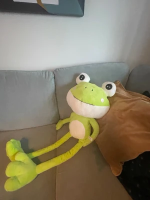 Stor grön groda mjukisdjur - Säljer ett stort mjukisdjur i form av en groda. Grodan är ljusgrön med vit mage och stora ögon med svarta pupiller. Den har långa armar och ben samt ett brett leende. Tillverkad i mjukt plyschmaterial, perfekt som dekoration eller kramkompis.