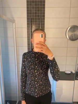 Svart blommig långärmad topp - Mysig svart långärmad topp med småblommigt mönster. Hög hals och rak passform, perfekt att styla med jeans eller kjol. Liten rund logga nertill. Superfin till både vardag och när du vill känna dig extra söt. Bara använd några gånger💞 Mått: längd: 53 cm (ej med polo-delen).  Bredd: 33 cm.  Stretchigt material💞 Om du tycker mönstret var fint men vill ha den i storlek 170, så säljer jag en HELT NY också🥰🥰😉