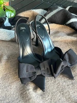 Svarta klackskor med rosett från Zara - Säljer ett par eleganta svarta pumps från Zara i storlek 38. Skorna har spetsig tå, hög klack och stora dekorativa rosetter framtill. Använda 1 gång till min student mottagning 