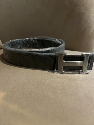 Svart H’er’m”e’s bälte med H-spänne - Stilrent svart bälte från H’er’m”e’ med H spänne. Bältet är tillverkat i skinn och har en elegant, enkel design som passar till många olika outfits. Perfekt accessoar för att lyfta din look.