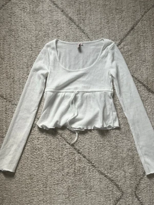 Vit ribbad topp med knyt i midjan - Supersöt vit långärmad topp med ribbad struktur och knytband i midjan ifrån Nelly. Den är i storlek XS och nästan aldrig använd🥰