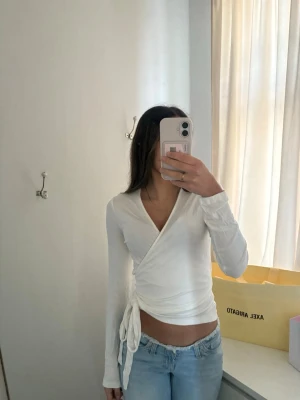 Vit omlottopp med knyt i sidan - Snygg vit omlottopp med lång ärm och knyt i sidan. Aldrig använd med prislappen kvar storlek S. (Säljer även jeansen i tidigare annons)