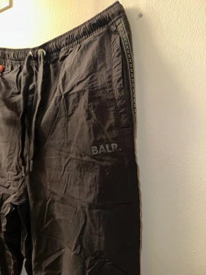 Svarta mjukisbyxor från BALR/PUMA - Svarta mjukisbyxor från BALR. med vita detaljer på benen och dragsko i midjan. Byxorna har resår vid benslut och en dekorativ söm längs sidorna. Perfekta för en avslappnad och sportig stil. Dem är special edition från Puma och ett samarbete med BALR