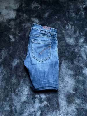Dondup George jeans - Snygga blå slim fit jeans från Dondup med lätt slitna detaljer och klassisk femficksdesign. Jeansen har modellen George och har knäppning framtill och en bakficka med dekorativ söm. Perfekta för en trendig och avslappnad look.