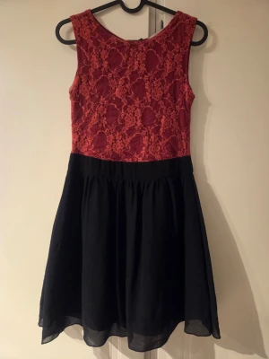 Svart och röd spetsklänning - Kort klänning med röd spets upptill och svart kjoldel. Ärmlös modell med rund halsringning och dragkedja i ryggen. Kjolen är lätt utställd och har ett tunt, svepande tyg. Perfekt för dig som vill ha en klänning med kontrasterande färger och fina spetsdetaljer.