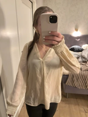 Krämvit blus med v-ringning - Säljer en krämvit blus med v-ringning och långa ärmar. Blusen har smockade muddar vid ärmslut och är tillverkad i ett tunt, lätt material som ger ett flowigt intryck. Perfekt för dig som gillar en enkel och stilren look. Säljer då den inte kommer till användning!❣️