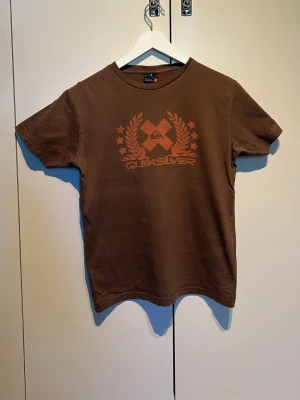 Brun Quiksilver t-shirt med tryck - Snygg brun t-shirt från Quiksilver med orange tryck framtill. Klassisk passform med rund hals och korta ärmar. Perfekt för dig som gillar streetwear och vill ha en enkel men stilren look.