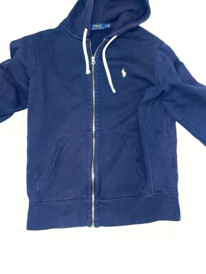 Marinblå hoodie från Polo Ralph Lauren - Snygg marinblå hoodie från Polo Ralph Lauren med dragkedja, vit dragsko och klassisk gul logga på bröstet. Hoodien har känguruficka framtill och är tillverkad i mjukt bomullsmaterial. Perfekt för en avslappnad och stilren look.