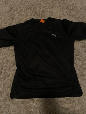 Svart tränings-tshirt från Puma storlek M - Snygg svart tränings-tshirt från Puma med diskret logga på bröstet. Tillverkad i ett lätt och snabbtorkande funktionsmaterial som passar perfekt till gymmet eller löprundan. Klassisk passform med korta ärmar och rund hals. Storlek m