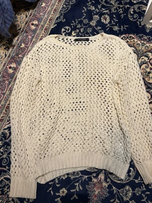 Vit hålstickad tröja - Säljer en vit hålstickad tröja med rund hals och ribbade muddar. Tröjan har ett luftigt, öppet stickat mönster och är perfekt för lager på lager. Materialet känns mjukt och stretchigt, troligen bomull eller bomullsblandning. Lång ärm och avslappnad passform.