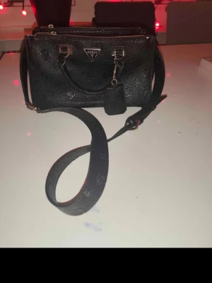 Svart handväska från Guess - Snygg svart handväska från Guess med präglat mönster och silverfärgade detaljer. Väskan har både handtag och justerbar axelrem samt flera fack med dragkedja. Perfekt för dig som vill ha en stilren och praktisk accessoar.