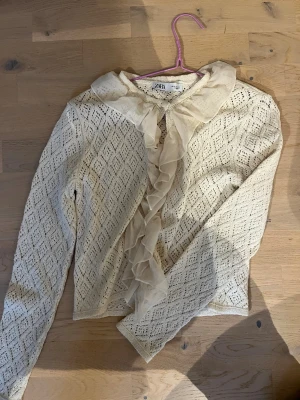 Vit spetskofta med volang från Zara - Snygg vit spetskofta från Zara med långärmad design och dekorativ volang framtill. Blusen har ett elegant mönster i spets och en mjuk, transparent volang längs knäppningen. Perfekt för dig som gillar romantisk och trendig stil.