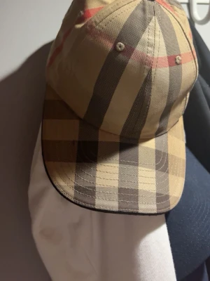 Beige keps med rutigt mönster - Snygg keps i klassiskt beige rutigt mönster med svarta, bruna och röda ränder. Kepsen har böjd skärm och justerbar baksida. Perfekt accessoar för en trendig streetstyle-look.