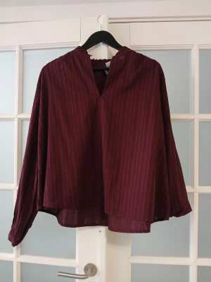 Vinröd blus med v-ringning from H&M - Snygg vinröd blus med subtila vertikala ränder och v-ringad hals. Blusen har långa, lösa ärmar med knapp vid ärmslut och är tillverkad i ett lätt, halvtransparent tyg som ger en luftig känsla. Perfekt att styla med jeans eller kjol.