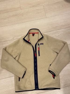 Beige patagonia fleece  - Säljer en beige fleecejacka från Patagonia med marinblå detaljer och dragkedjor. Jackan har hel dragkedja framtill, två sidofickor och en bröstficka med röd dragkedja. Perfekt för lager-på-lager, fleecen är i storleken xxl i barn vilken motsvarar ungefär S skulle jag säga. Hör av er vid frågor eller funderingar priset går att diskutera!