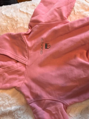 Rosa hoodie från GANT med logga - Säljer en snygg rosa hoodie från GANT med broderad logga och sköld på bröstet. Hoodien har klassisk huva och en stor magficka framtill. Tillverkad i mjukt bomullsmaterial som är perfekt för chill dagar. Enkel och stilren design som funkar till det mesta.