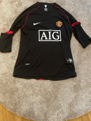 Manchester United svart matchtröja Nike - Snygg svart Manchester United matchtröja från Nike med röd detaljsöm och klubbmärke på bröstet. AIG-logga på magen och Nike Swoosh på höger sida. Tillverkad i funktionsmaterial med Dri-FIT-teknologi. Perfekt för fotboll eller träning.
