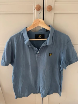 Blå pikétröja från Lyle & Scott - Säljer en klassisk blå pikétröja från Lyle & Scott med gul logga på bröstet. Tröjan har korta ärmar, krage och knappar framtill. Perfekt för en avslappnad och stilren look.