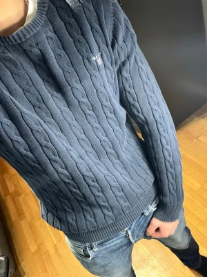 Blå kabelstickad tröja från GANT - Säljer en blå kabelstickad tröja från GANT med rund halsringning och diskret logga på bröstet. Tröjan har ribbade muddar vid ärmslut och nederkant, och är tillverkad i ett mjukt stickat material som passar perfekt till jeans.